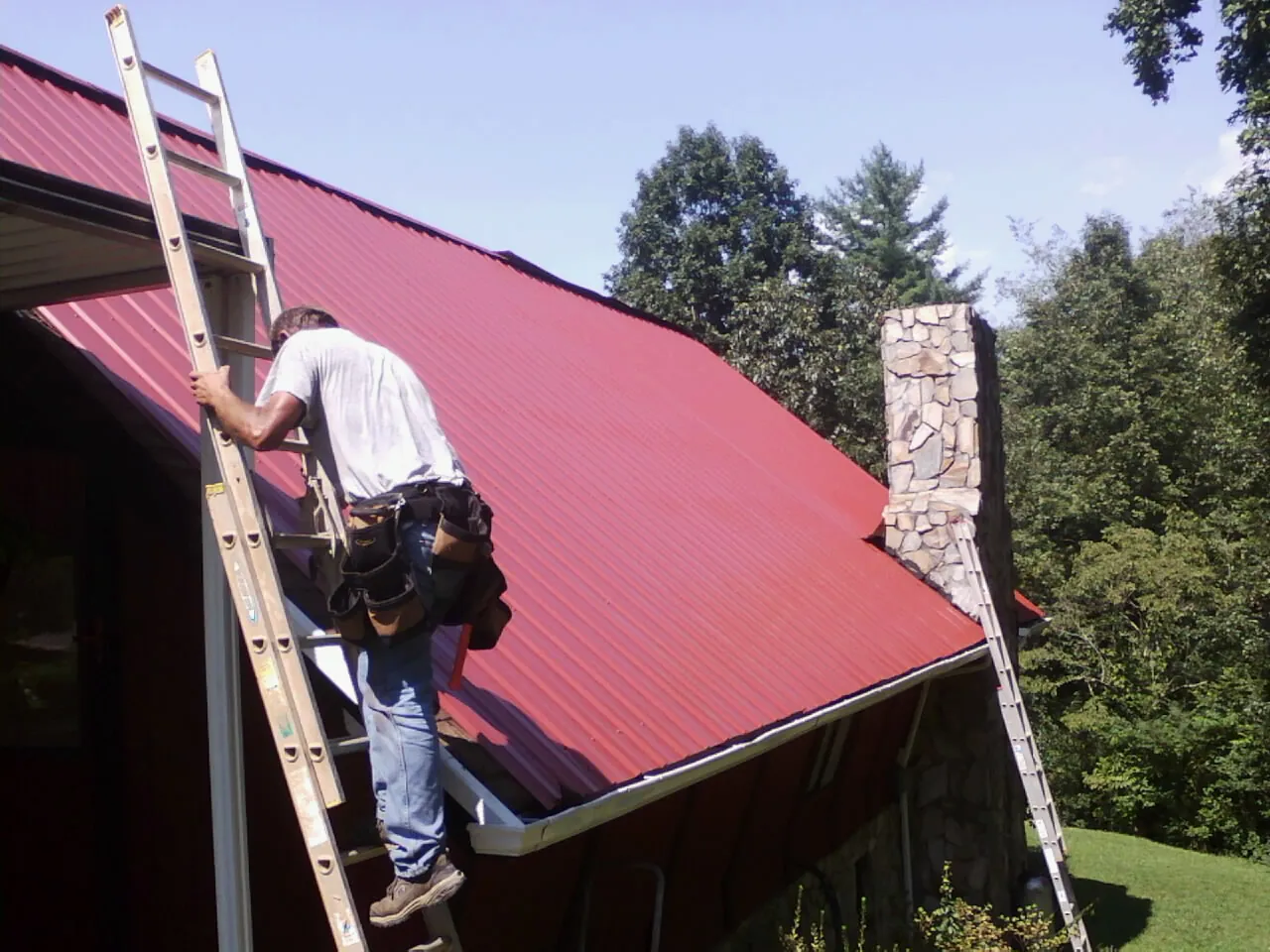 Roofer on a ladder beside a red metal roof and stone chimney — Soffit & Fascia Repair by Roofing Brothers in South Ozone Park